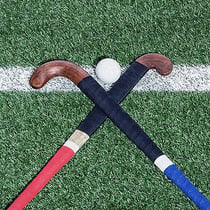 Two diagonally crossed field hockey sticks lying on a playing field. In between is a field hockey ball on one side.